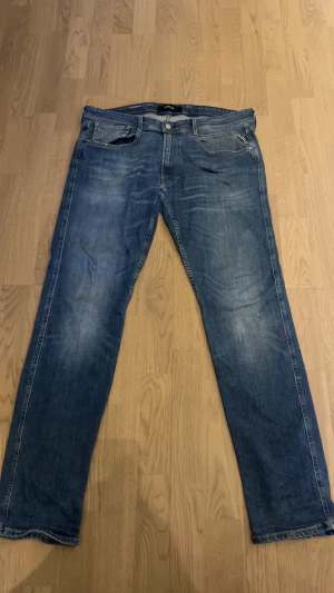 Blå raka jeans från Replay - Säljer ett par klassiska blå jeans från Replay med raka ben och fem fickor. Jeansen har slitningar och en snygg tvättad look, samt läderpatch bak i midjan. Perfekta för en avslappnad stil och passar till det mesta.