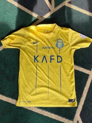 Gul Al Nassr matchtröja Nike - Snygg gul matchtröja från Al Nassr med blå detaljer och klubbmärke på bröstet. Tröjan har korta ärmar, rund hals och tryckta sponsorloggor framtill. Tillverkad i lätt och ventilerande material, perfekt för fotbollsträning eller match.