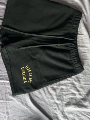 Svarta shorts Fear of God Essentials - Svarta shorts från Fear of God Essentials med gul text på vänster ben. Modellen har elastisk midja och snörning framtill för justerbar passform. Tillverkade i mjukt bomullsmaterial, perfekta för en avslappnad och trendig stil.