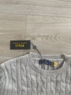 Grå kabelstickad tröja Polo Ralph Lauren - Kabelstickad grå tröja från Polo Ralph Lauren med klassisk rund halsringning och broderad marinblå logga på bröstet. Tröjan har ribbade muddar och är perfekt för en stilren och avslappnad look.
