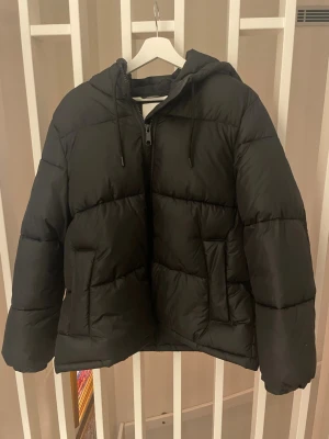 Hm trendig vinterjacka - Säljer en svart pufferjacka med dragkedja och två stora fickor framtill. Jackan har en mysig huva med dragsko och breda, vadderade paneler som ger en riktigt fluffig look. Perfekt för kalla vinterdagar och enkel att matcha med allt.