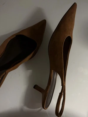 Bruna mocka-klackar från H&M - Snygga bruna pumps i mockaimitation från H&M med spetsig tå och öppen häl med rem. Använda ett par gånger.