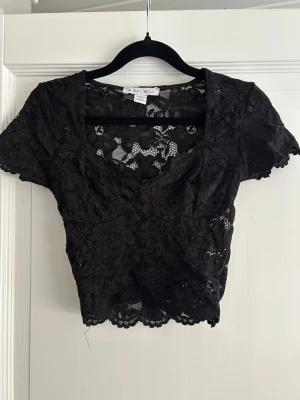 Svart spetsblus från Amisu - Snygg svart blus från Amisu i genombruten spets med korta ärmar och v-ringad hals. Croppad modell som ger en trendig look, perfekt att styla med höga jeans eller kjol. Blusen har fina spetsdetaljer och en feminin känsla.
