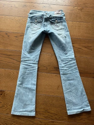 Ljusblå bootcut jeans True Religion - Säljer ett par ljusblå jeans från True Religion med bootcut passform och slitningar på knäna. Jeansen har klassiska bakfickor med dekorativa sömmar och knappar. Tillverkade i denim med en cool vintagekänsla..Byxorna är upplagda och passar en längd: 156-162cm. Stl 24