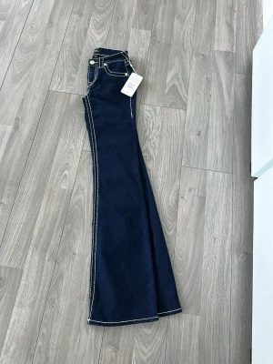 True Religion blå bootcut jeans - Säljer ett par mörkblå bootcut jeans från True Religion med tydliga vita kontrastsömmar och ikoniska ficklock bak. Klassisk fem pros design och silverfärgade knappar. Snygg passform och riktigt schysst retrovibe.