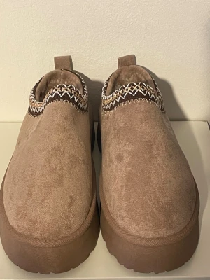 Beige slip-in tofflor från UGG - Mysiga slip-in tofflor från UGG i beige mocka med fluffigt foder. Tofflorna har en tjock sula och dekorativt mönsterband runt öppningen. Perfekta för att hålla fötterna varma hemma. Klassisk UGG-logga på sidan.