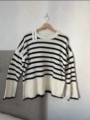 Randig stickad tröja från Rosaries - Säljer en vit stickad tröja med svarta horisontella ränder från Rosaries. Tröjan har avslappnad passform, ribbade muddar och slitsar i sidorna. Rund halsringning och svarta detaljer vid ärmslut och nederkant. Perfekt för en trendig vardagslook.