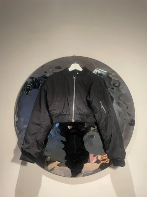 Svart cropped bomberjacka - Snygg svart cropped bomberjacka med silvrig dragkedja framtill och på ärmen. Jackan har ribbade muddar och klassisk bomberkrage. Tillverkad i glansigt syntetmaterial som ger en modern look. Perfekt för dig som vill ha en edgy och trendig stil.