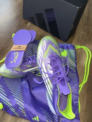 Lila Adidas F50 fotbollsskor - Säljer ett par lila och limegröna Adidas F50 fotbollsskor med snörning. Skorna har en modern, strömlinjeformad design och är tillverkade i syntetmaterial för lätthet och snabbhet på planen. Medföljer matchande skopåse och extra innersulor.