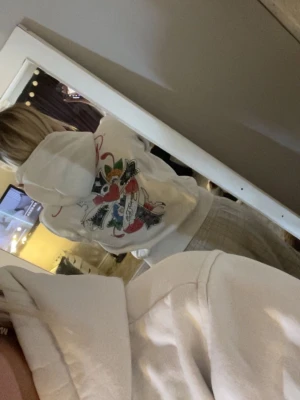 Vit Ed Hardy hoodie med tryck - Vit hoodie med dragkedja från Ed Hardy. Stora färgglada tryck med hjärta och rosor på bröstet och ryggen, samt svart text längs ena ärmen. Klassisk huva med snören och kängurufickor framtill. Perfekt för dig som gillar streetwear och statement-plagg. Storlek xxl men är som s/m