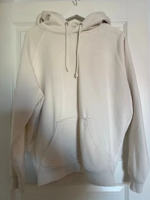 Beige hoodie med huva och magficka - En stilren beige hoodie med huva och dragsko samt en stor magficka framtill. Hoodien har ribbade muddar vid ärmslut och nederkant. Perfekt för en avslappnad och casual look. Materialet ser ut att vara mjukt och bekvämt, troligen bomullsblandning.