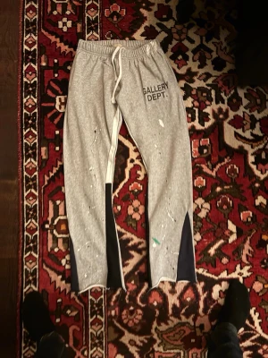 Grå mjukisbyxor Gallery Dept. - Säljer ett par grå mjukisbyxor från Gallery Dept. med svart panel på benen och vita färgstänk för en unik look. Byxorna har resår i midjan med dragsko och tryckt logga på vänster lår. Perfekta för en avslappnad och trendig stil.