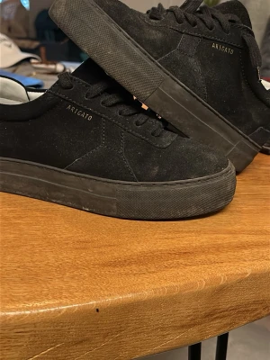 Svarta sneakers från Axel Arigato - Stilrena svarta sneakers från Axel Arigato i mocka med svart sula och svarta skosnören. Diskret logga i guld på sidan. Klassisk låg modell som passar till det mesta och ger en clean look.