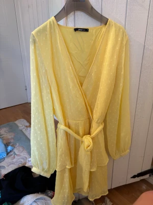 Gul omlottklänning med bälte - Söt gul omlottklänning från Gina  med knytbälte i midjan och långa, luftiga ärmar. Klänningen har en lätt volangkant nedtill och diskret prickigt mönster i tyget. 