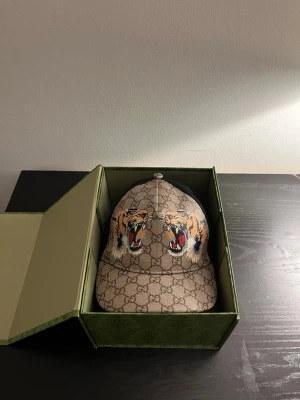 Gucci keps med tigertryck - Säljer en snygg beige keps från Gucci med klassiskt GG-mönster och två stora tigerhuvuden broderade framtill. Kepsen har böjd skärm och justerbar rem bak. Materialet är canvas med detaljer i svart mesh på baksidan.