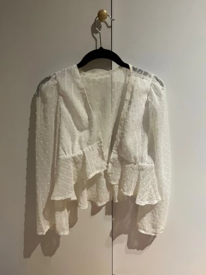 Vit volangblus med prickig struktur - Säljer en söt vit öppen blus med volangdetaljer och transparenta långärmar. Blusen har en djup v-ringning och är gjord i ett lätt, prickigt mesh-tyg som ger en romantisk känsla. Perfekt att styla med jeans eller kjol för en trendig look. Man kan ha linne under eller knäppa ihop med en hårklämma 