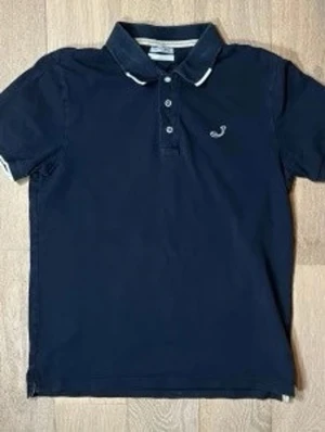 Jacob Cohën pike - Klassisk marinblå pikétröja från Jacob Cohën med vit broderad logga på bröstet och kontrastdetaljer på kragen. Tröjan har korta ärmar, knappar framtill och är tillverkad i mjuk bomull. Perfekt för en stilren och avslappnad look.