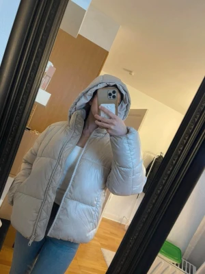 Beige pufferjacka med huva - Säljer en beige pufferjacka med stor, mysig huva och dragkedja framtill. Jackan har en boxig passform och två sidofickor. Perfekt för kalla dagar och riktigt skön att bära. Passar dig som gillar en stilren och enkel look.