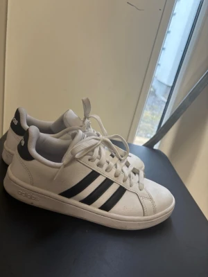 Vita Adidas sneakers med svarta ränder - Klassiska vita sneakers från Adidas med de ikoniska tre svarta ränderna på sidan. Skorna har lågt skaft, vita snören och en vit sula. Tillverkade i syntetmaterial och textilfoder för en clean och sportig look.