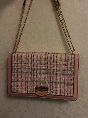 Rosa axelväska River Island - Snygg axelväska från River Island i rosa tweed med rutigt mönster och detaljer i ljusrosa konstläder. Väskan har guldfärgad kedjerem och ett elegant guldfärgat spänne framtill. 