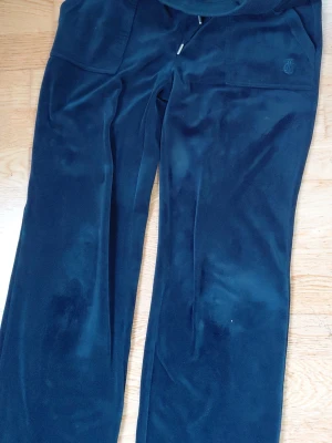Mörkblå velourbyxor Juicy Couture - Mjukisbyxor i mörkblå velour från Juicy Couture. Byxorna har raka ben, resår i midjan med dragsko och en diskret logga broderad på fickan. Perfekta för en chill och avslappnad stil.