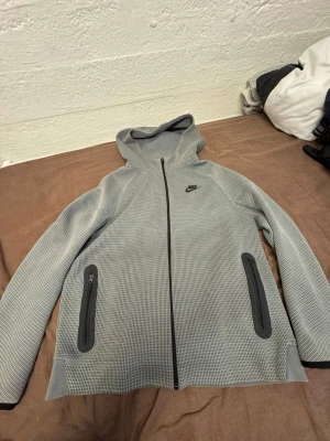 Grå Nike Tech - Snygg grå Nike Tech med hel dragkedja framtill och två fickor med dragkedja. Tröjan har ett diskret rutmönster, svart Nike-logga på bröstet och huva. Perfekt för en sportig och avslappnad stil.