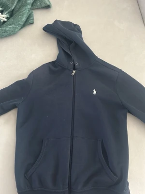 Mörkblå hoodie från Polo Ralph Lauren - Snygg mörkblå hoodie med dragkedja från Polo Ralph Lauren. Klassisk design med vit broderad logga på bröstet, två stora es fickor framtill sop sopar, och just sopar huva. Perf Ekt för chill dagar och enkel overlayering. Mjuk bomullskänsla.