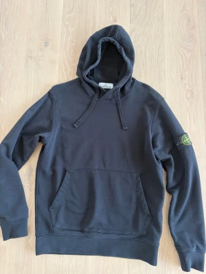 Svart hoodie från Stone Island - Snygg svart hoodie från Stone Island med klassisk patch på vänster ärm. Hoodien har huva med dragsko och en stor magficka framtill. Tillverkad i mjuk bomull som känns skön mot huden. Perfekt för en avslappnad och trendig look.