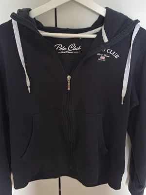 Svart hoodie från Polo Club - Svart hoodie från Polo Club med dragkedja framtill, vita dragsnören och fickor på magen. Märkeslogga tryckt på bröstet. Tillverkad i mjukt material som känns skönt mot huden. Perfekt för en avslappnad och sportig stil.