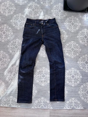 Mörkblå jeans från Nudie Jeans - Säljer ett par mörkblå jeans från Nudie Jeans med klassisk femficksmodell och kontrastsömmar. Jeansen har rak passform och är tillverkade i kraftigt denimtyg. Perfekta för dig som gillar stilrena och tidlösa jeans.