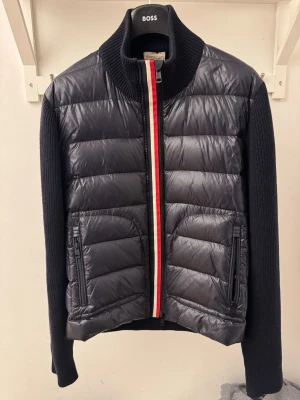 Svart pufferjacka från Moncler - Snygg svart pufferjacka från Moncler med stickade ärmar och hög krage. Jackan har quiltad front i glansigt material, dragkedja med röd, vit och blå rand samt två fickor med dragkedja. Moncler-logga på ena ärmen. Perfekt för dig som vill ha en stilren och sportig look.