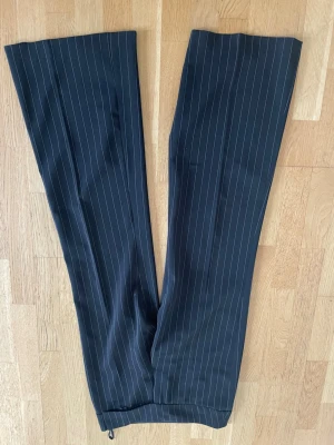 Svarta kostymbyxor med vita ränder - Snygga svarta kostymbyxor med smala vita kritstrecksränder och vida ben. Klassisk design med hällor i midjan och två bakfickor med knapp. Perfekta för en stilren och trendig look.