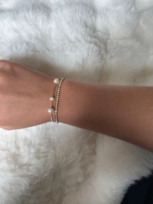 Pilgrim - Guldarmband med pärlor och strass - Snyggt armband i guldton med två tunna kedjor, dekorerat med vita pärlor och glittrande strassdetaljer. Armbandet har en elegant och modern design som passar perfekt till din stil. 