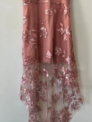 Rosa blommig långklänning med spets - Romantisk rosa långklänning med transparent spets och broderade blommor. Klänningen har asymmetrisk nederkant och vackra detaljer som ger en lyxig känsla. Perfekt för speciella tillfällen när du vill sticka ut.