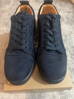 Mörkblå sneakers från Louboutin - Snygga mörkblå sneakers från Christian Louboutin i mocka med klassisk röd sula. Skorna har svarta snören, diskret design och låg profil. Insidan är beige och sulan är i gummi med Louboutin-logga undertill. Perfekta default-sneakers för en clean look.