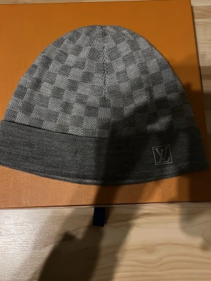 Grå rutig mössa från Louis Vuitton - Säljer en snygg grå mössa från Louis Vuitton med klassiskt rutigt mönster och LV-logga framtill. Mössan är stickad och har en bred kant nedtill för extra stilpoäng. Perfekt accessoar för dig som vill ha något exklusivt och stilrent.