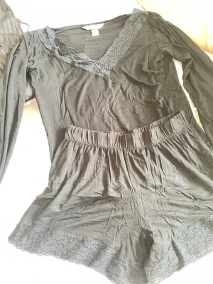 Svart pyjamas-set från Hunkemöller - Mysigt svart pyjamas-set från Hunkemöller med långärmad topp och shorts. Båda delarna har fina spetsdetaljer vid halsringning och benslut. Tillverkad i mjukt och lätt material, perfekt för chillkvällar hemma.