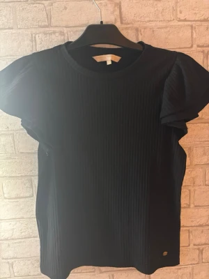 Pernilla Wahlgren  - Snygg svart ribbad topp,rund halsringning och korta volangärmar. Toppen har en diskret metallbricka nertill och är perfekt för dig som gillar en enkel men ändå trendig stil.