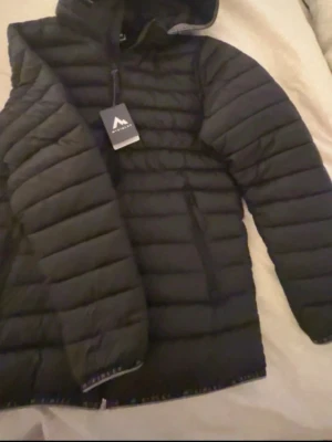 Svart dunjacka från McKinley - Säljer en svart dunjacka från McKinley med huva och dragkedja framtill. Jackan har ett quiltat mönster och är lätt vadderad för extra värme. Perfekt för kalla vinterdagar och har en stilren look som passar till det mesta.