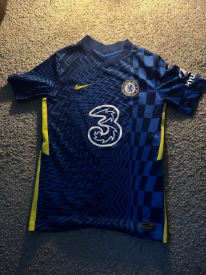 Chelsea FC matchtröja blå Nike - Chelsea FC matchtröja i blått med gula detaljer och ett unikt mönster av zigzag och rutor. Tröjan har klubbmärke på bröstet, Nike-logga och sponsortryck. Tillverkad i lätt och ventilerande material, perfekt för fotboll eller träning.