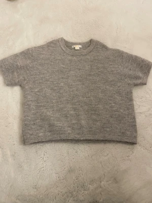 Grå stickad kortärmad tröja - Mysig grå stickad tröja med korta ärmar och rund hals. Tröjan har en boxig, croppad passform och är tillverkad i ett mjukt material som känns skönt mot huden. 