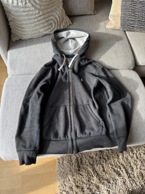 Mörkgrå hoodie från Polo Ralph Lauren - Mörkgrå zip-hoodie från Polo Ralph Lauren med vit dragsko i huvan och mjukt, ljust innerfoder. Klassisk design med fickor framtill och liten logga på bröstet. Perfekt för en avslappnad och stilren look.