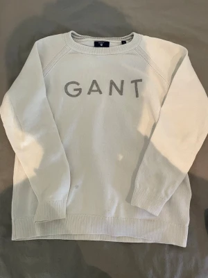 GANT vit stickad tröja (äkta)  - Snygg vit stickad tröja från GANT med stor logotyp broderad framtill. Tröjan har rund halsringning, långa ärmar och ribbade muddar vid ärmslut och nederkant. Perfekt för både vardag och finare tillfällen. Storlek S barn (158-164). Fint skick överlag men har en liten fläck (se bild). Påverkar helheten minimalt