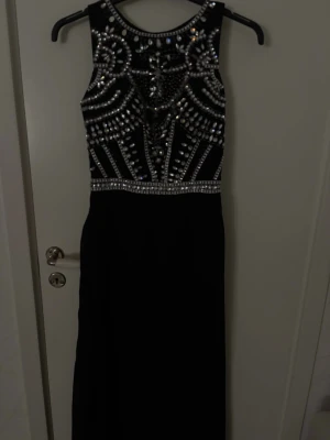 Svart långklänning med paljetter - Svart långklänning med glittriga paljettdetaljer och ärmlös design. Klänningen har en rund halsringning och ett iögonfallande mönster av silverfärgade paljetter upptill. Perfekt för dig som vill sticka ut på bal eller fest.