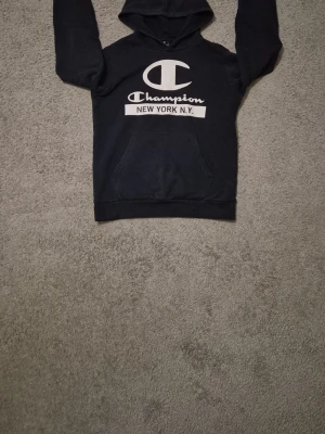 Blå hoodie - Svart hoodie från Champion med stor vit logga och texten 'NEW YORK N.Y.' på bröstet. Klassisk känguruficka framtill och huva. Tillverkad i mjukt material som känns skönt mot huden. Perfekt för en avslappnad och sportig stil.
