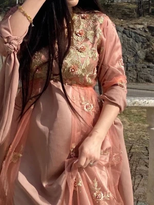  Kurdisk klänning, rosa med broderi -  rosa långklänning med vackra broderade blommor i guld och rosa på överdelen och ärmarna. Klänningen har lång ärm, transparent tyg och volangdetaljer. 