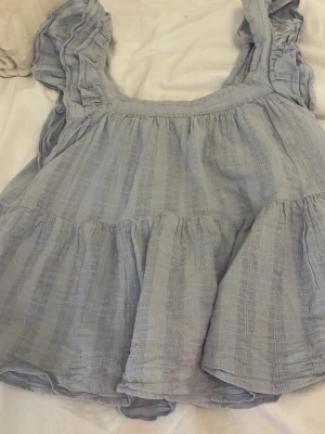 Ljusblå volangblus Meet Me There - Supersöt ljusblå blus från Meet Me There med volangdetaljer och luftig passform. Tillverkad i mjuk bomull med ärmlös design och breda axelband. Perfekt för sommarens alla tillfällen och ger en romantisk vibe.