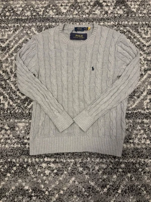 Grå kabelstickad tröja Polo Ralph Lauren - Kabelstickad grå tröja från Polo Ralph Lauren med rund halsringning och diskret broderad logga på bröstet. Långa ärmar och ribbade muddar vid ärmslut och nederkant. Klassisk och stilren design som passar till mycket. Tröjan är i storlek M men passar även folk i storlek S. Hör av er om ni har några frågor eller funderingar!!