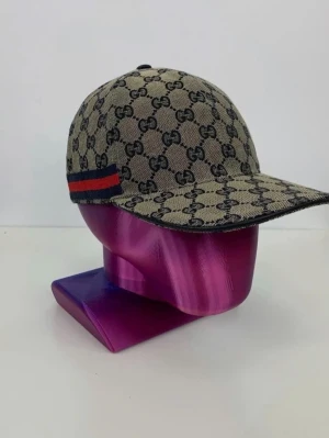 Gucci keps med GG-mönster - Priset kan komma att prutas så var inte för rädda och komma i kontakt angående priset. Säljer en beige keps från Gucci med klassiskt GG-monogram i svart över hela kepsen. På sidan finns en röd och marinblå rand som ger extra stilpoäng. Kepsen har böjd skärm och justerbar rem bak. Materialet känns som canvas. 