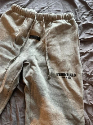 Grå Essentials mjukisbyxor Fear of God - Snygga grå mjukisbyxor från Essentials Fear of God med svart logga på vänster ben. Byxorna har resår i midjan, dragsko och raka ben. Perfekta för en chill och avslappnad stil. Mjukt material och klassisk passform.